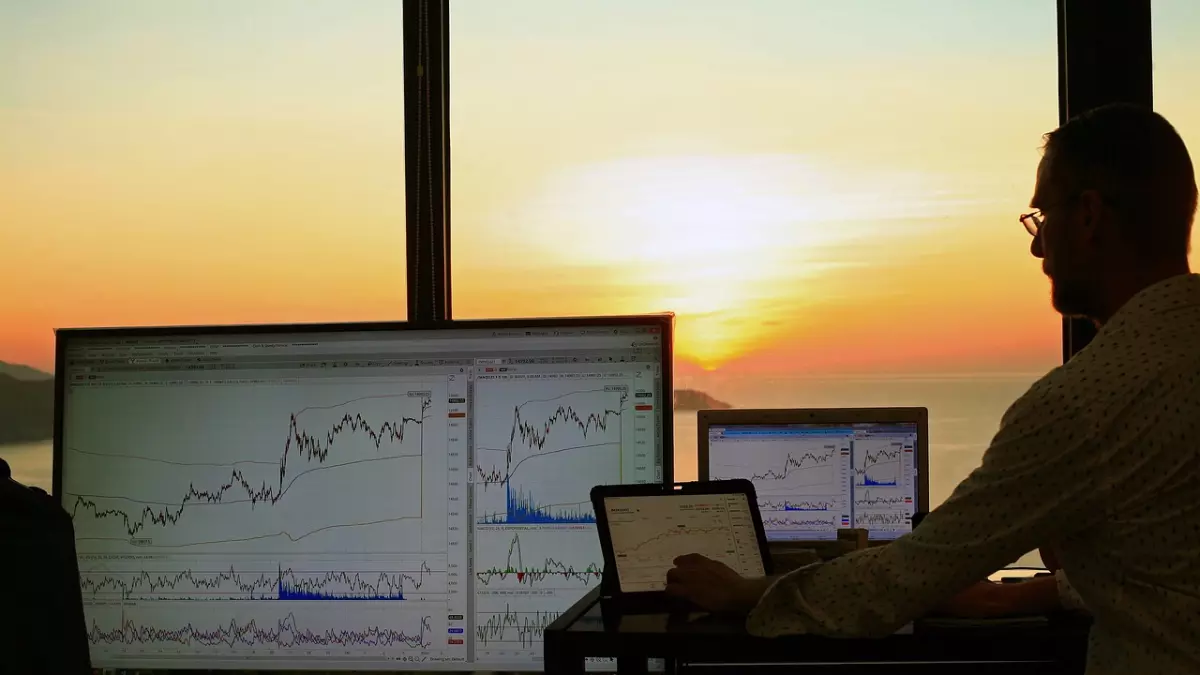 A man sits at a desk facing multiple monitors displaying financial charts. The sun sets in the background, casting a warm glow. 